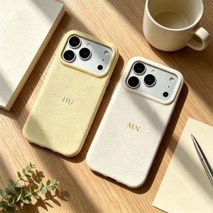 May include: Two smartphone cases with a textured finish, one in pale yellow with the letters HU and one in off-white with the letters MN. Each case features a raised camera cutout with three circular lens openings and a flash aperture on the back.
