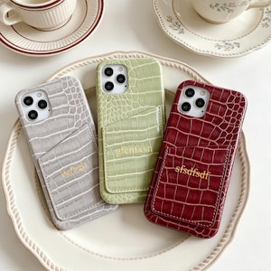 May include: Three phone cases in gray, green, and burgundy, each with a crocodile skin texture and a cardholder pocket. The gray case has the text "hdhck", the green case has "gfenfxsn", and the burgundy case has "sfsdfsdf".