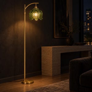 May include: A floor lamp with a slender brass-toned metal stand and a circular base. The lamp features a curved arm holding a translucent, scalloped green glass shade that emits a warm, glowing light from the bulb housed inside the fixture.