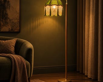 May include: A floor lamp with a slender gold-toned metal stand and a circular base. The lamp features a dome-shaped green fabric shade adorned with decorative hanging fringe trim and tassels, emitting a warm, soft glow from within.