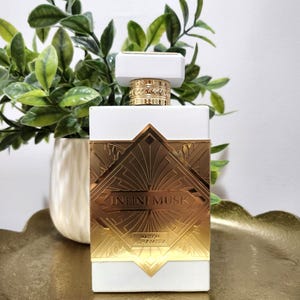 May include: A rectangular white perfume bottle with a gold-toned metallic plate featuring geometric art deco patterns. The text "INFINI MUSK" and "MAISON ALHAMBRA" are embossed on the gold plate. The bottle has a white cap and a gold collar.