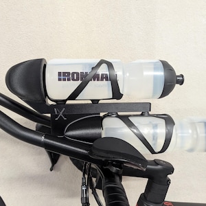 May include: A black bicycle hydration system mounted between the handlebars, featuring two translucent white water bottles with black caps and cages. The top bottle displays the black text IRONMAN. The mount is black metal with a small IX logo.