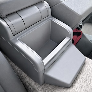 May include: A light grey plastic automotive centre console storage compartment with an open rectangular bin and a hinged lid mechanism, featuring a smooth matte finish and a stepped design integrated into the vehicle interior seating area.