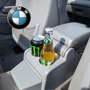 Può includere: Una console centrale per auto in plastica grigia con due portabicchieri contenenti una lattina di bevanda energetica Monster nera e una bottiglia di birra Saku Ice in vetro trasparente. Il logo BMW è visibile nell'angolo in alto a sinistra.
