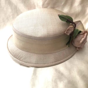 May include: A light beige hat with a wide brim and a delicate pink rose accent. The hat is made of a woven fabric and has a satin ribbon trim.