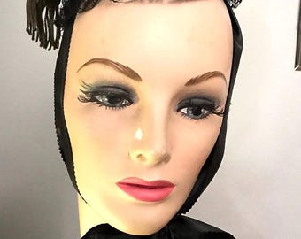 Puede incluir: Un sombrero de terciopelo negro con un gran lazo y un borde de encaje. El sombrero se lleva en un maniquí con un velo de encaje negro y un collar de perlas.