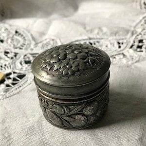 May include: A silver trinket box with an ornate floral design. The box has a hinged lid and is decorated with raised flowers and leaves.