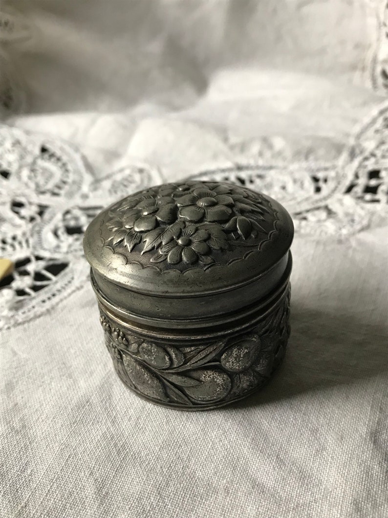 May include: A silver trinket box with an ornate floral design. The box has a hinged lid and is decorated with raised flowers and leaves.