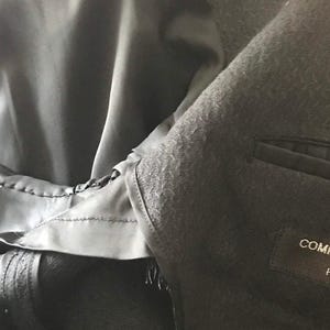 Pode incluir: Close-up de um blazer preto com tecido texturizado. O interior do blazer é forrado com um tecido preto liso. O rótulo no blazer diz "COMME des GARCONS HOMME PLUS".