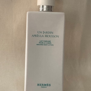 May include: A white bottle of Hermes Un Jardin Après La Mousson perfumed body lotion. The bottle has a white cap and a green label with the product name and description.