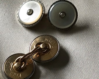 May include: A pair of silver cufflinks with a mother of pearl design. The cufflinks have a small pearl in the center of each. The cufflinks are on a white fabric background.