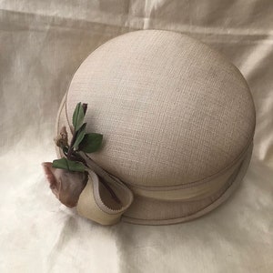 May include: A beige straw hat with a wide brim and a small, light brown flower and green leaves on the side.