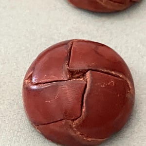 Könnte beinhalten: Nahaufnahme von zwei runden, rotbraunen Knöpfen. Die Knöpfe haben ein segmentiertes Design, mit sichtbaren Nähten, die ein kreuzartiges Muster bilden. Die Knöpfe scheinen aus Leder oder einem ähnlichen Material zu bestehen.