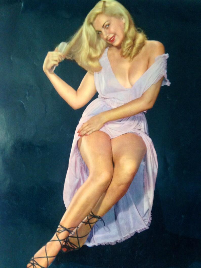 May include: A woman with long blonde hair is wearing a light purple dress with a low neckline. She is sitting with her legs crossed and wearing black strapped heels.