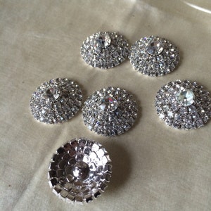 May include: Six silver-toned, round, rhinestone embellishments with a clear crystal center. The embellishments are arranged in a semi-circle on a light beige surface.