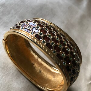 以下が含まれることがあります： 茶色と緑のエナメル模様の金製のバングルブレスレット。エナメルは幾何学模様で、小さな緑色のラインストーンが飾られています。