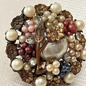 Puede incluir: Un broche vintage con un diseño floral que presenta una perla central rodeada de perlas más pequeñas, pedrería y cuentas de colores en tonos rosa, rojo y azul.