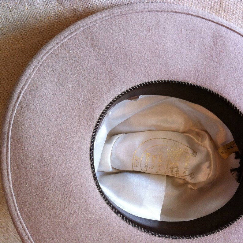 May include: A beige felt fedora hat with a brown leather band. The hat has a white satin lining with a gold horseshoe logo and the text "Five Church Duck by the Sea" printed on it.