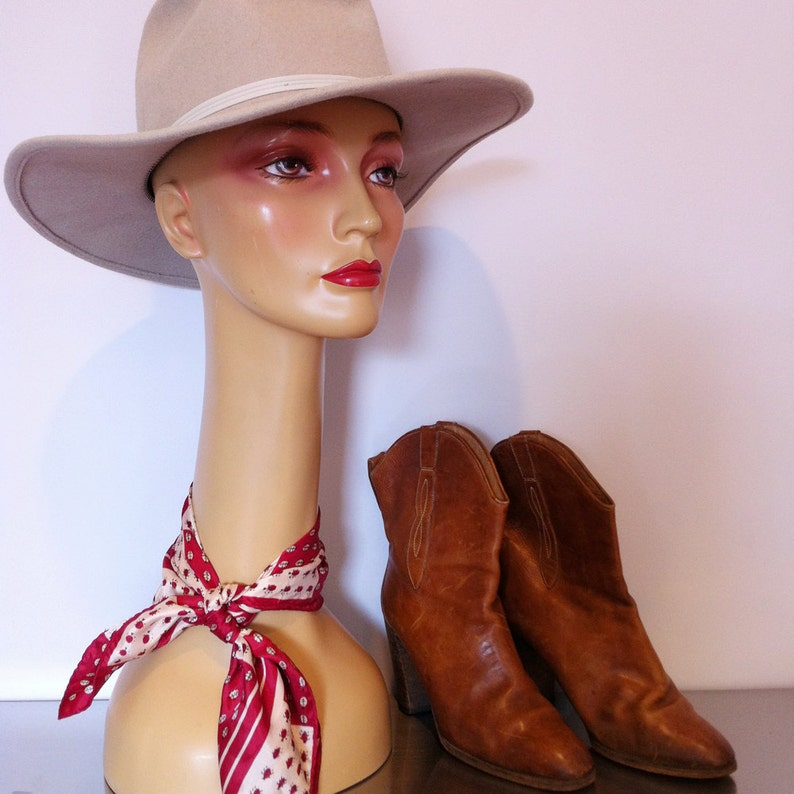 May include: A mannequin wearing a beige cowboy hat, a red and white patterned scarf, and a pair of brown leather cowboy boots.
