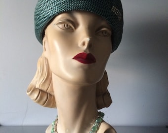 May include: A green and white straw hat with a decorative pin on the side. A mannequin is wearing the hat and a green and silver necklace.