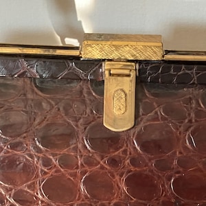 May include: Close-up of a vintage brown handbag with a textured, crocodile-like pattern. The bag features a gold-colored clasp and trim. The clasp is rectangular with a textured surface and a gold-colored latch.