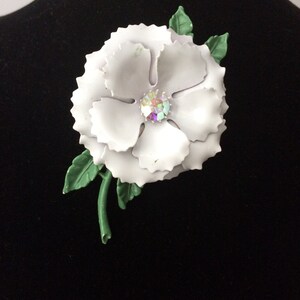 以下が含まれることがあります： 緑色の茎と葉が付いた白いエナメル製のフラワーブローチ。花の中央には大きな虹色のラインストーンが埋め込まれています。