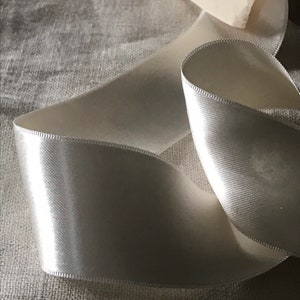 May include: A white satin ribbon, folded and lying on a textured fabric surface.