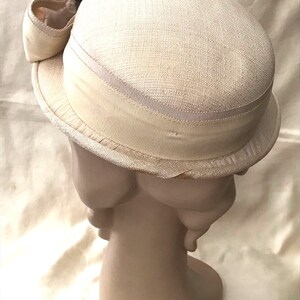 May include: A white straw hat with a wide brim and a white satin ribbon around the crown. The hat is decorated with a pink flower and green leaves.