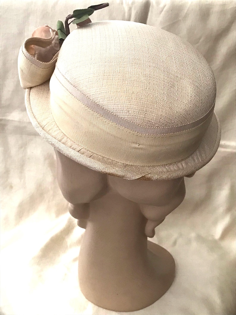 May include: A white straw hat with a wide brim and a white satin ribbon around the crown. The hat is decorated with a pink flower and green leaves.