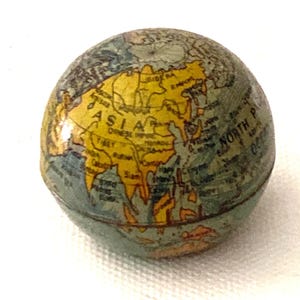 Puede incluir: Un pequeño globo terráqueo vintage con un mapa del mundo detallado. Los continentes están coloreados en tonos amarillos, verdes y azules. La palabra "ASIA" se muestra de forma destacada. El globo es esférico y parece estar hecho de papel o material similar.