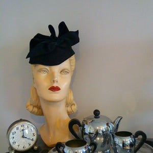 Könnte beinhalten: Ein Vintage-Mannequin-Kopf mit einem schwarzen Samthut mit Schleife. Das Mannequin trägt roten Lippenstift. Ein silbernes Teekannen- und Milchkännchen-Set befindet sich auf einem silbernen Tablett vor dem Mannequin.