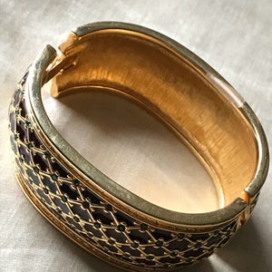 以下が含まれることがあります： 濃い茶色のエナメルと緑色のアクセントが施された、金色のバングルブレスレット。ブレスレットは幾何学的なパターンで、片側が開いています。