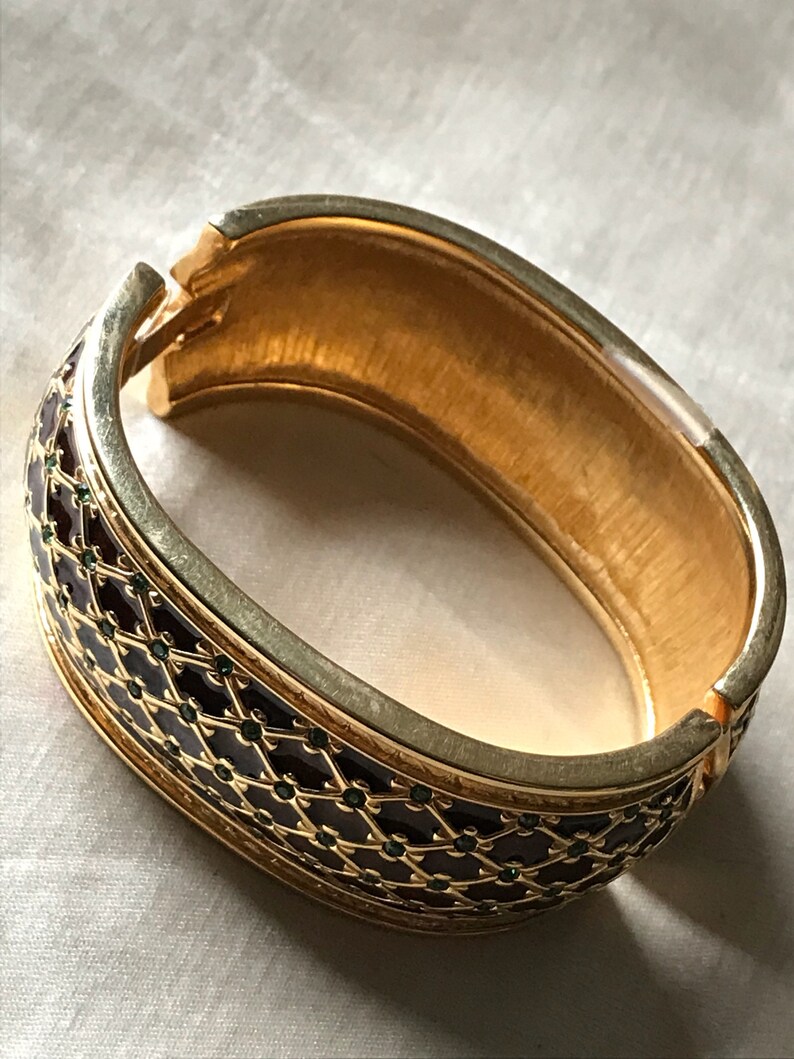 以下が含まれることがあります： 濃い茶色のエナメルと緑色のアクセントが施された、金色のバングルブレスレット。ブレスレットは幾何学的なパターンで、片側が開いています。
