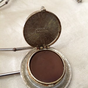 May include: A silver locket necklace with a circular pendant. The pendant opens to show a dark brown interior. The necklace features a chain of small silver beads and a bar-shaped chain. The locket is marked with the word "estee".