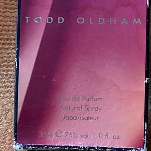 Pode incluir: Uma caixa rosa e prateada com o texto "TODD OLDHAM" impresso na frente. A caixa também inclui o texto "Eau de Parfum, Natural Spray, Vaporisateur, 30ml e 76% vol. 1.0 fl. oz."