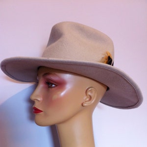 May include: A beige felt fedora hat with a small feather accent. The hat is worn on a mannequin's head.