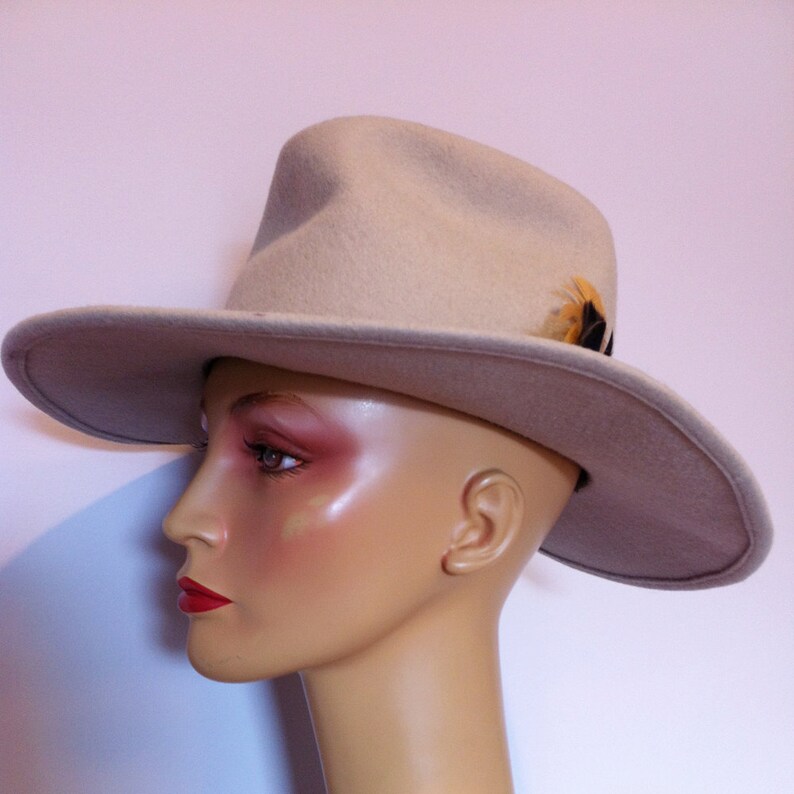 May include: A beige felt fedora hat with a small feather accent. The hat is worn on a mannequin's head.