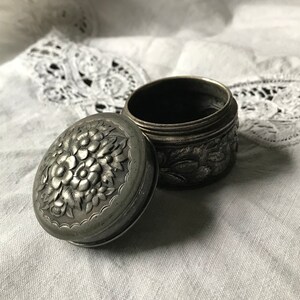 May include: A silver trinket box with an ornate floral design. The lid is open and the box is empty.