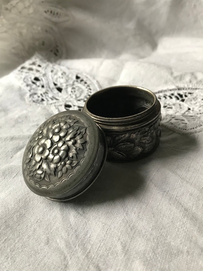 May include: A silver trinket box with an ornate floral design. The lid is open and the box is empty.