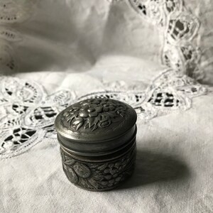 May include: A small, silver, round trinket box with an ornate floral design. The box has a hinged lid and is decorated with raised flowers and leaves.
