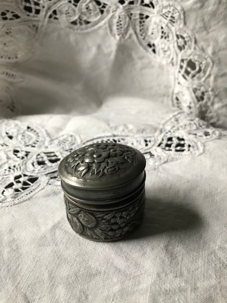 May include: A small, silver, round trinket box with an ornate floral design. The box has a hinged lid and is decorated with raised flowers and leaves.