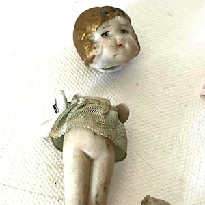 Puede incluir: Una muñeca vintage con la cabeza y el cuerpo separados. La cabeza de la muñeca está pintada con pelo dorado y una boca roja. El cuerpo lleva una falda verde. Un pequeño trozo de la muñeca está separado.