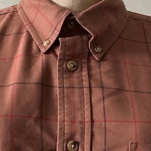Puede incluir: Una camisa de botones marrón con un patrón de cuadros formado por líneas finas rojas y azul oscuro. La camisa tiene un cuello puntiagudo con botones en las puntas, una tapeta central de botones y un bolsillo visible en el lado derecho.