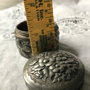 May include: A silver, round, embossed trinket box with a floral design. The box is approximately 2 inches in diameter. The box is sitting on a white fabric surface.