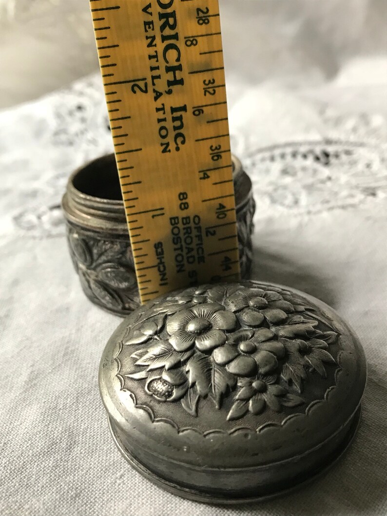 May include: A silver, round, embossed trinket box with a floral design. The box is approximately 2 inches in diameter. The box is sitting on a white fabric surface.