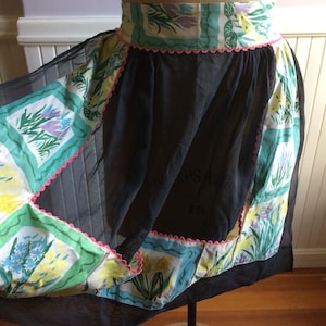 Könnte beinhalten: Eine vintage schwarze Netzschürze mit einem floralen Muster am Rand. Der Druck zeigt gelbe, grüne, blaue und violette Blumen. Die Schürze hat einen rosa gewellten Saum.