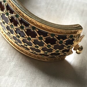 以下が含まれることがあります： 濃い茶色のエナメル模様と緑色の宝石が付いた金のバングルブレスレット。ブレスレットは、詳細で複雑なデザインです。
