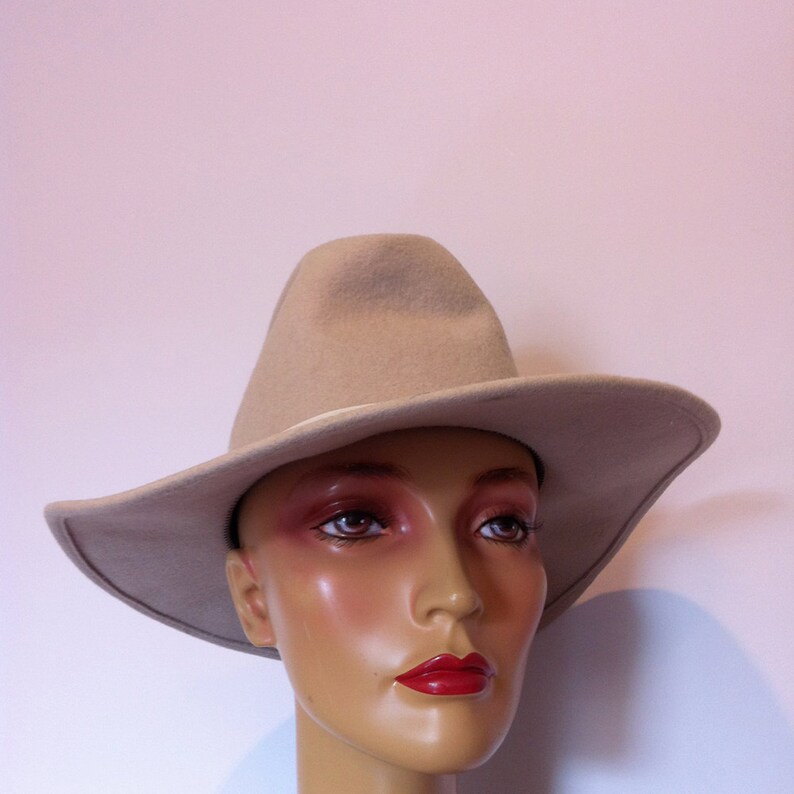 May include: A beige felt cowboy hat with a wide brim. The hat is on a mannequin head with neutral makeup and red lipstick.