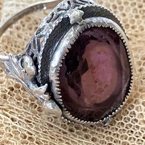 Könnte beinhalten: Ein silberner Ring mit einem filigranen Design und einem großen, runden, violetten Edelstein.