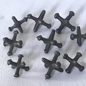 Pode incluir: Dez jacks de metal preto, também conhecidos como ossos dos dedos, estão espalhados sobre uma superfície branca. Os jacks têm a forma de uma estrela de quatro pontas.